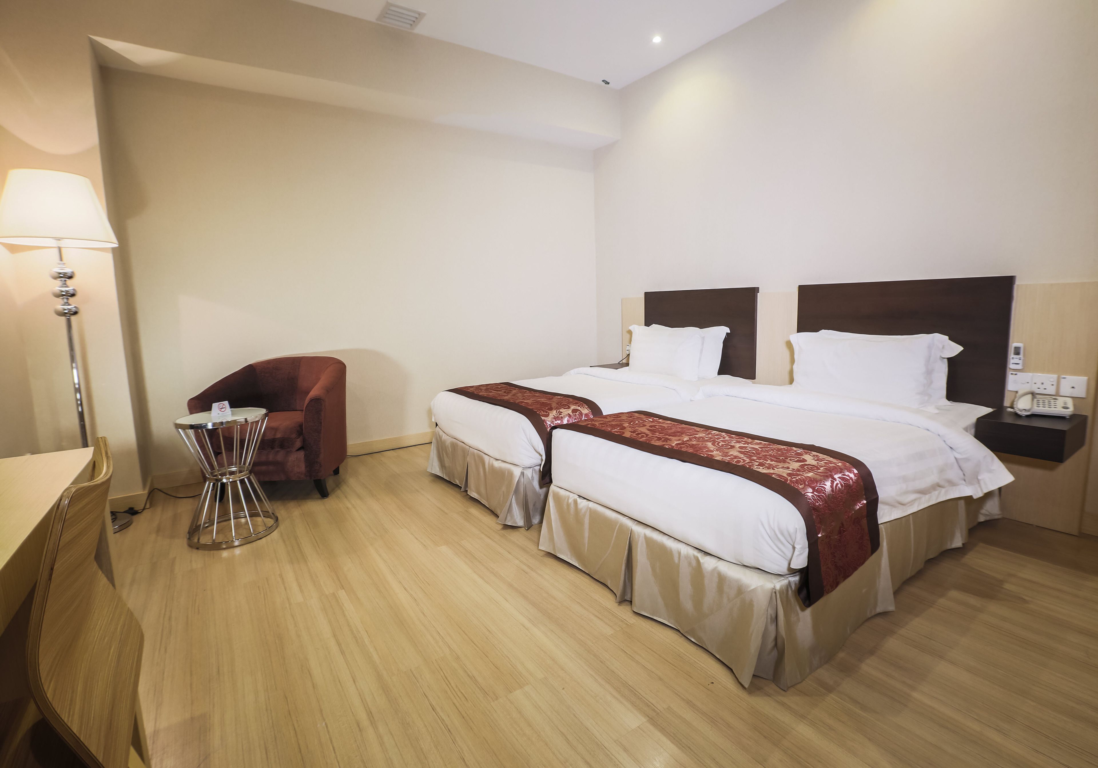 A hotel room with two beds, a desk, a chair, a lamp, and a bedside table.