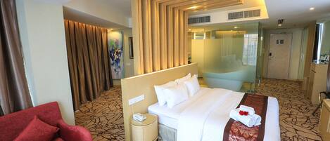 A hotel room with a large bed, a red armchair, a bedside table with a phone, and a glass partition wall.
