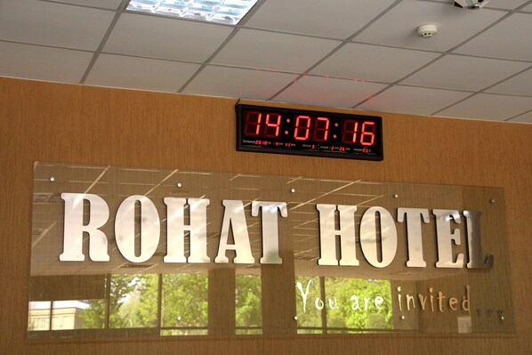 Un reloj digital muestra las 14:07:16 sobre un letrero que dice 'ROHAT HOTEL' con el texto 'Eres invitado' debajo.