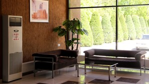 A modern waiting area with a wooden wall, a framed cityscape picture, a large window with a view of neatly trimmed hedges, and a parked car outside. - Rohat Hotel (Tashkent)