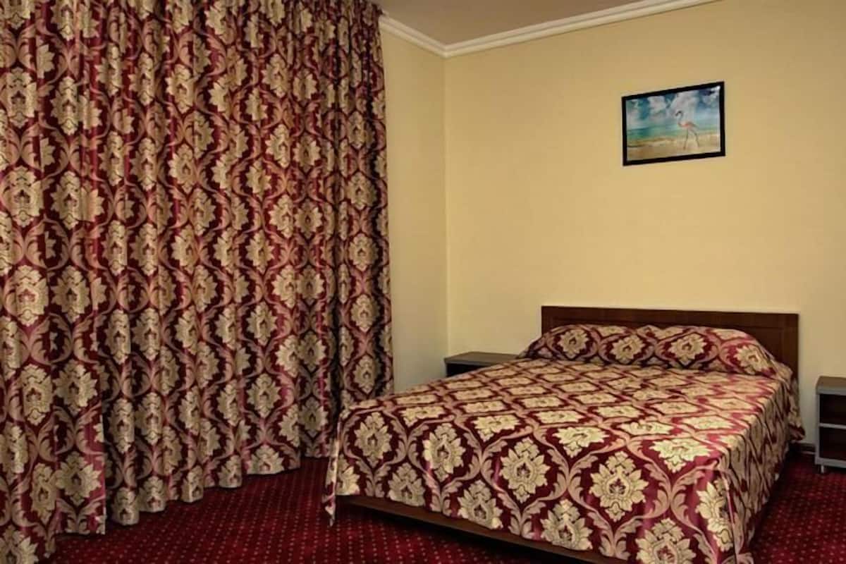 A bedroom with a patterned bedspread, a nightstand, a bedside table, and a painting on the wall.