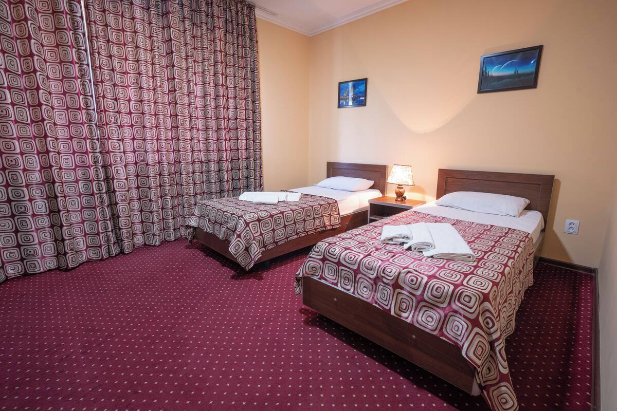 A hotel room with two beds, a patterned curtain, a night lamp, and two wall-mounted pictures.