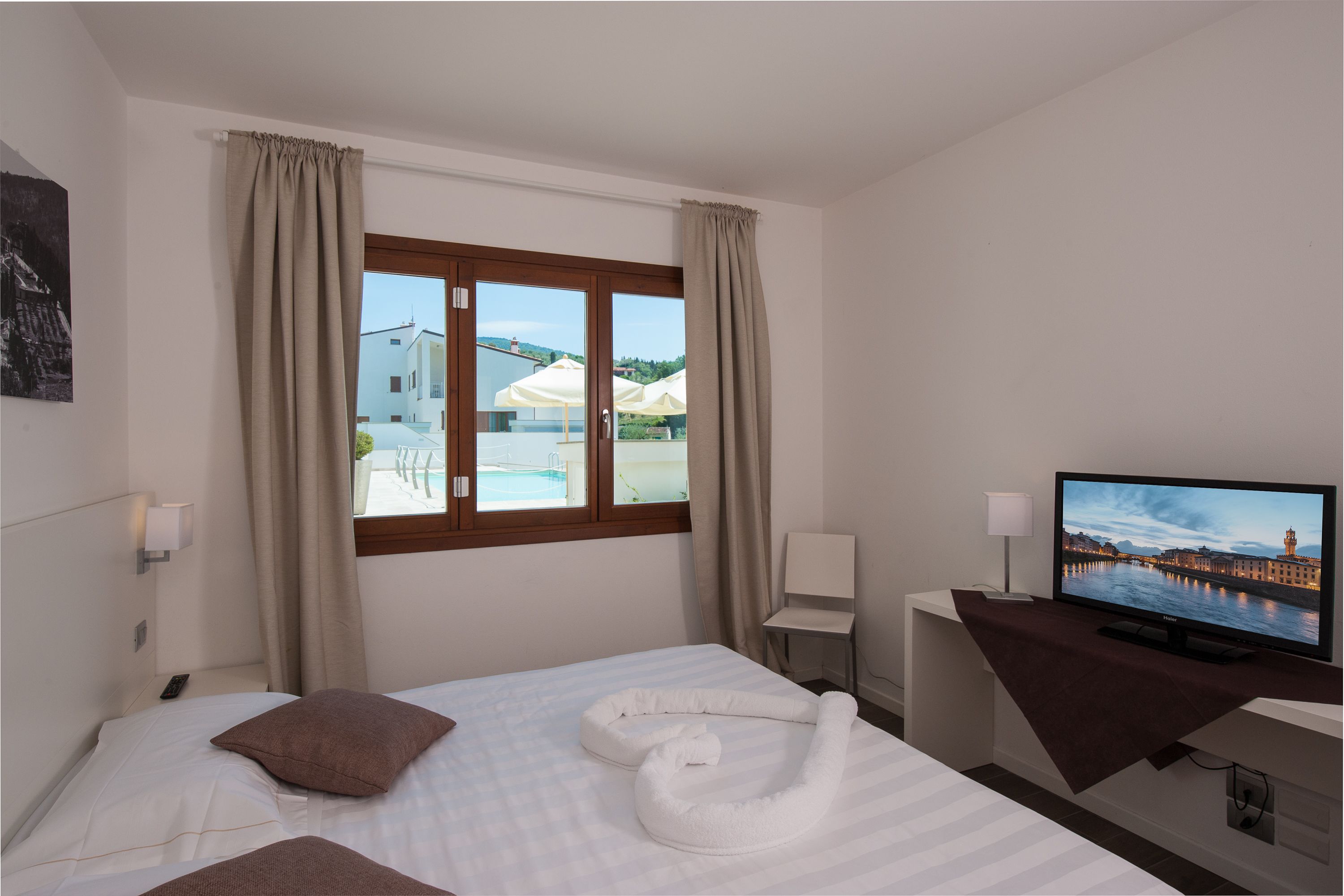 Ein Hotelzimmer mit Bett, Fernseher, Fenster mit Vorhängen und Blick auf einen Pool und Gebäude.