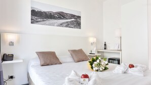 Et hotelværelse med en pænt redt seng, en telefon ved sengebordet, en lampe, et lille bord med en flaske og et glas, samt et indrammet billede på væggen.