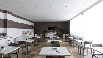 A modern dining area with white tables and chairs, a kitchen in the background, and large windows.