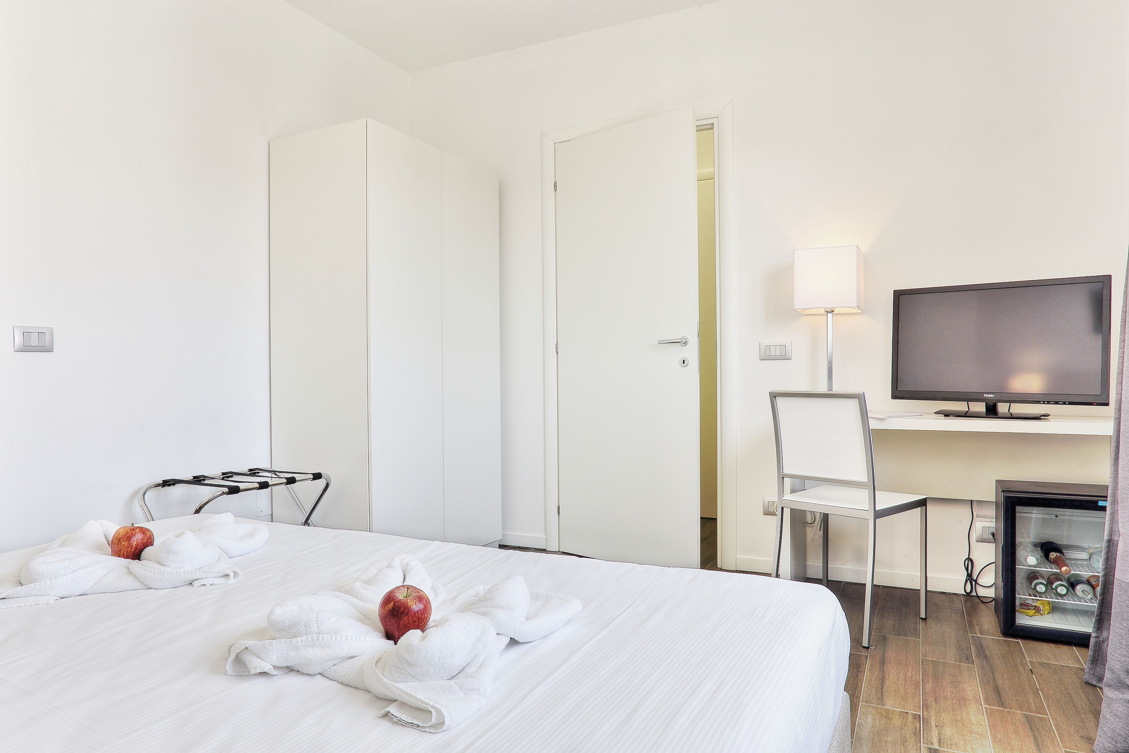 A hotel room with a bed, a desk with a television, a chair, and a minibar.