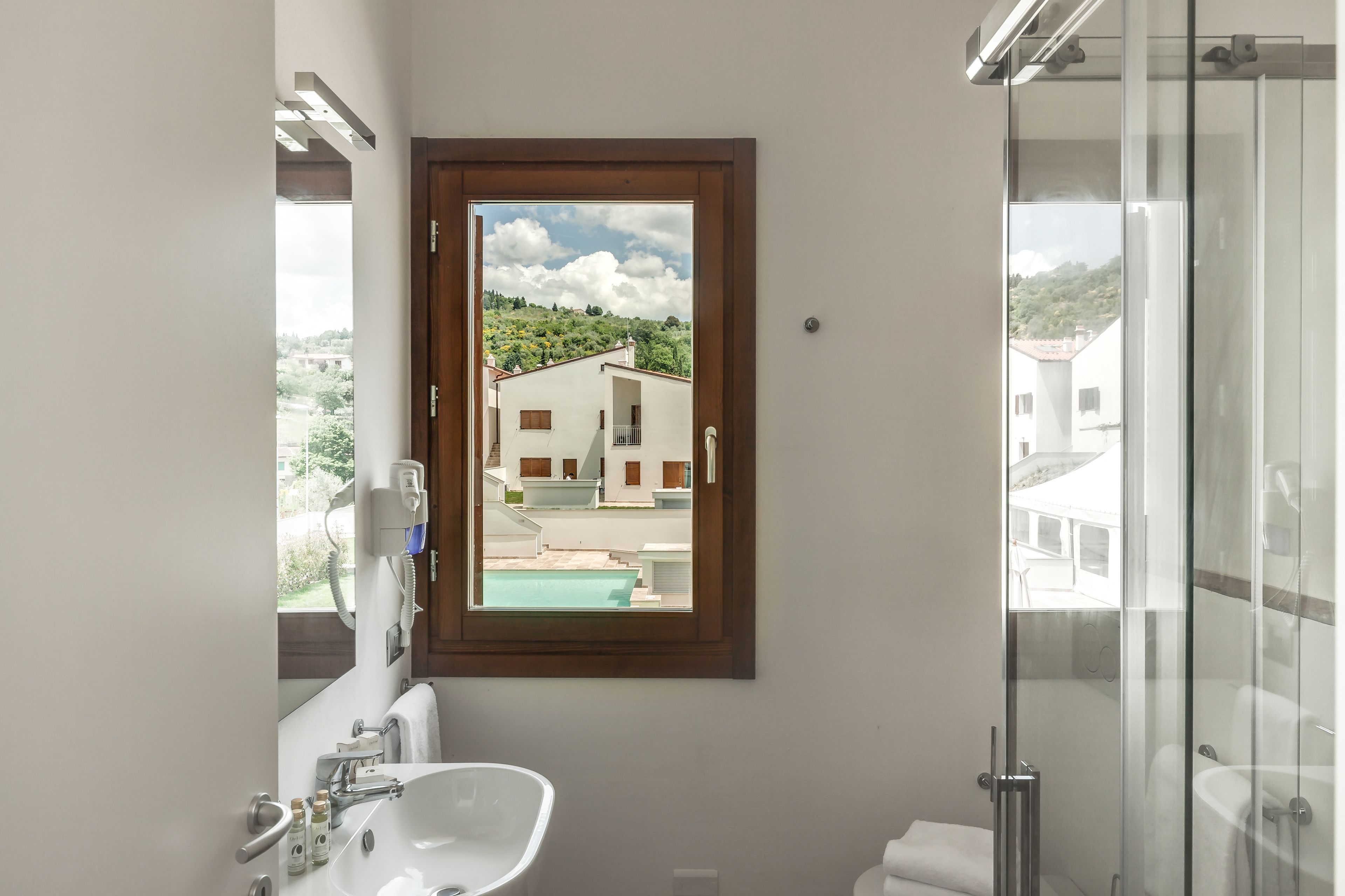 Ein Badezimmer mit Waschbecken, Spiegel und Dusche. Durch ein Fenster ist ein Blick auf Gebäude und einen Pool möglich.