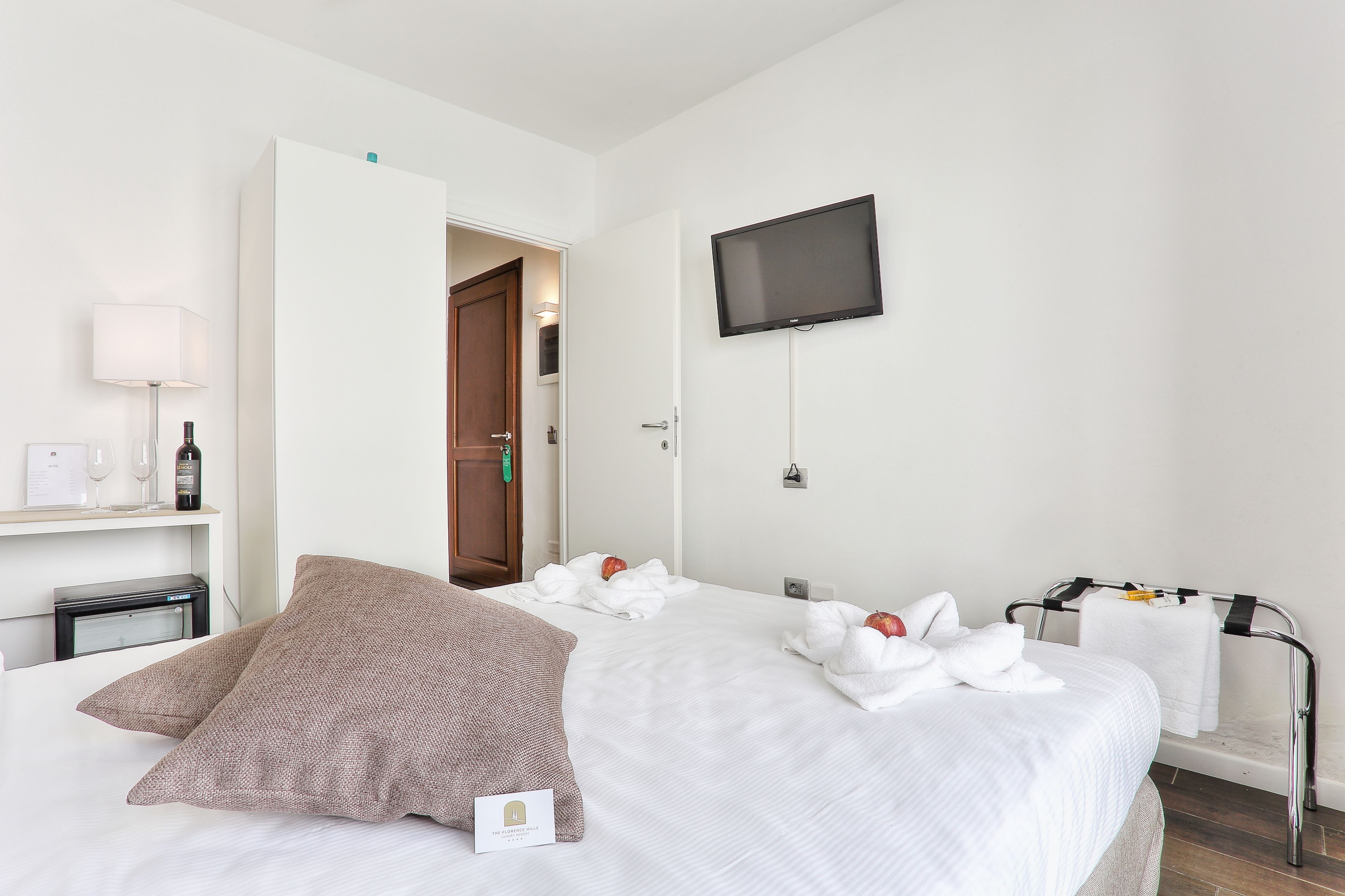 Habitación de hotel con dos camas, un televisor montado en la pared, una mesita de noche con lámpara y una puerta que da a otra habitación.