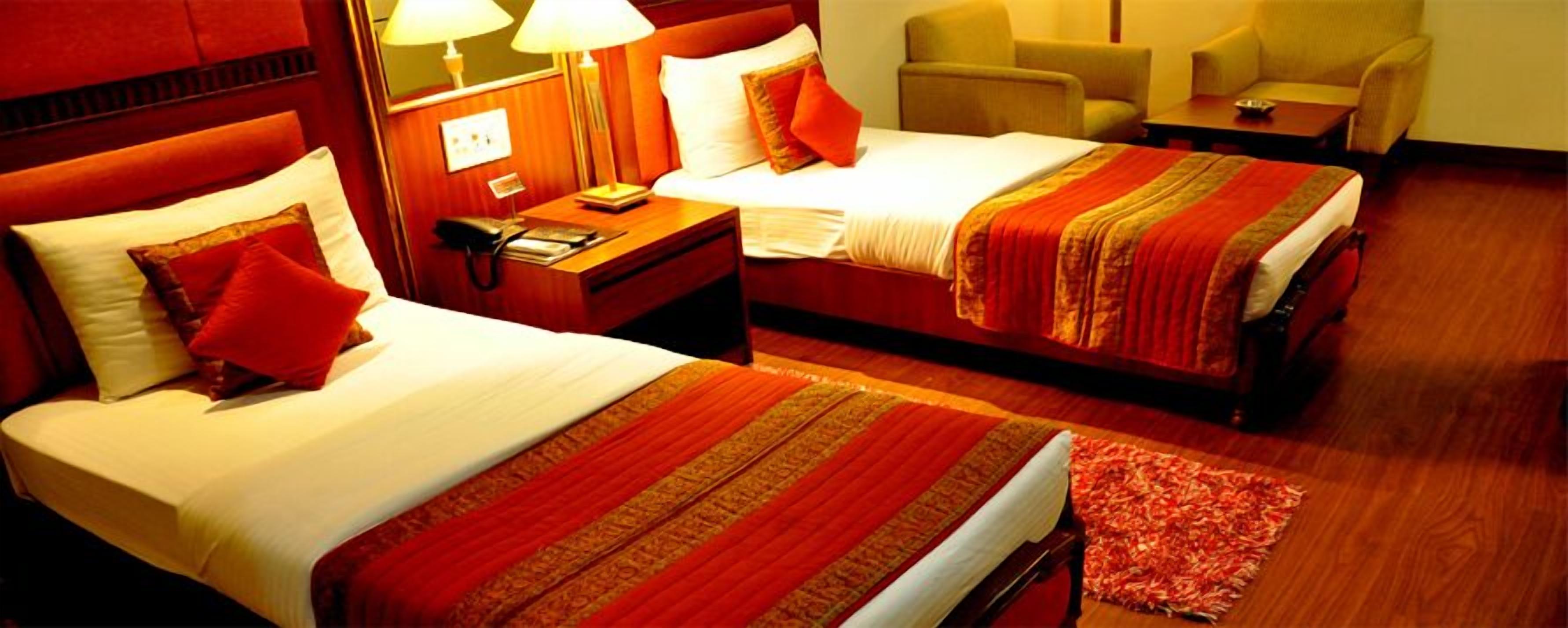 A hotel room with two beds, a nightstand, a lamp, a chair, and a small table.
