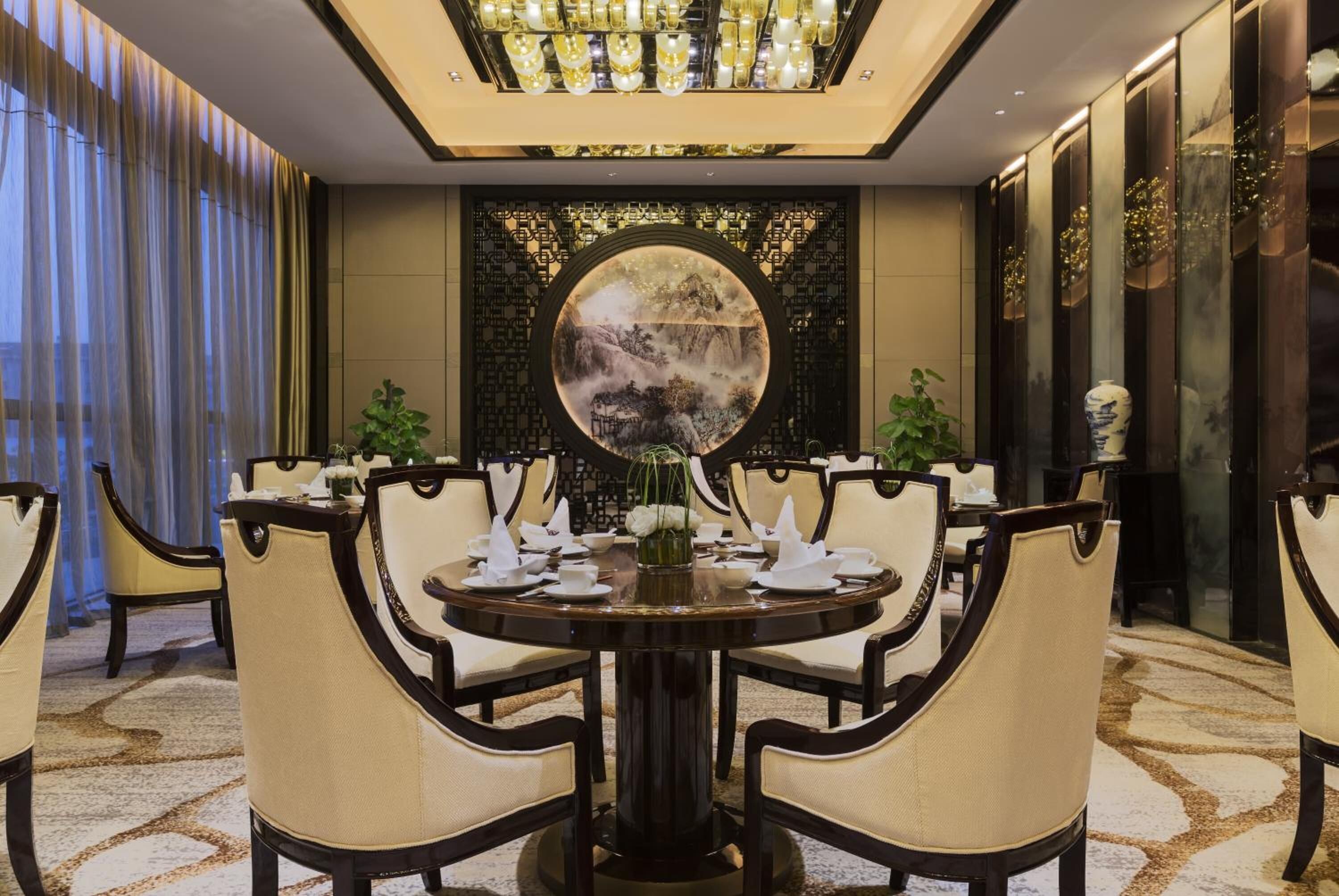 Un comedor con una mesa redonda preparada para seis personas, rodeada de sillas tapizadas, un jarrón decorativo y un espejo redondo montado en la pared.