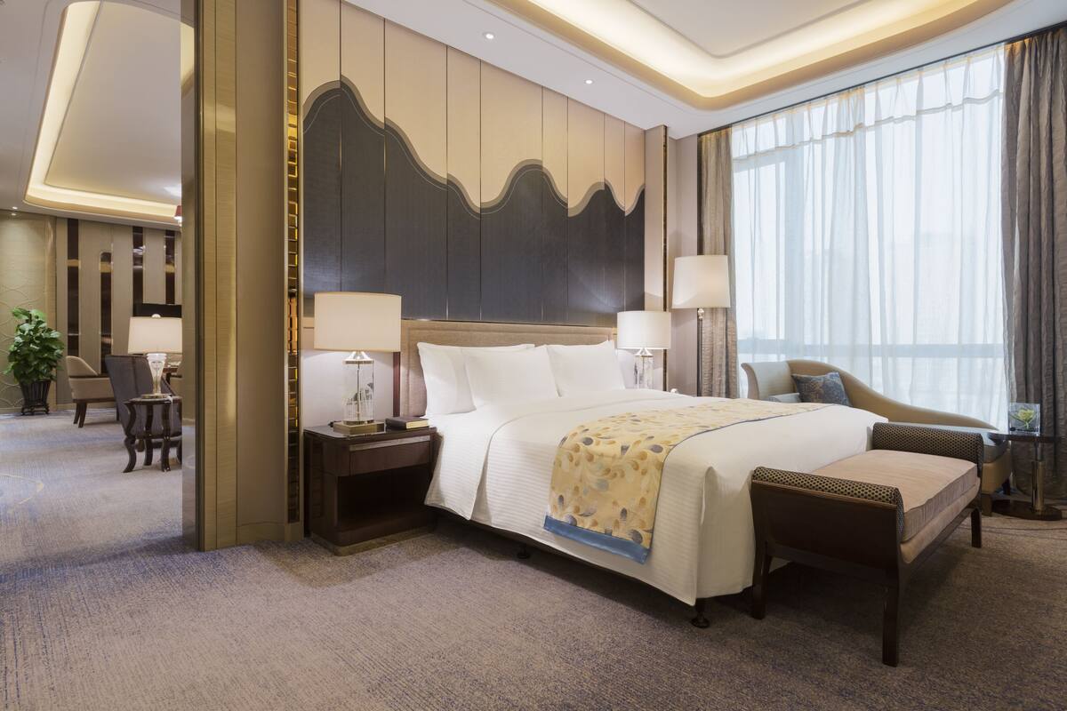 A hotel room with a large bed, two bedside tables, a chair, a desk, and a large window with curtains.