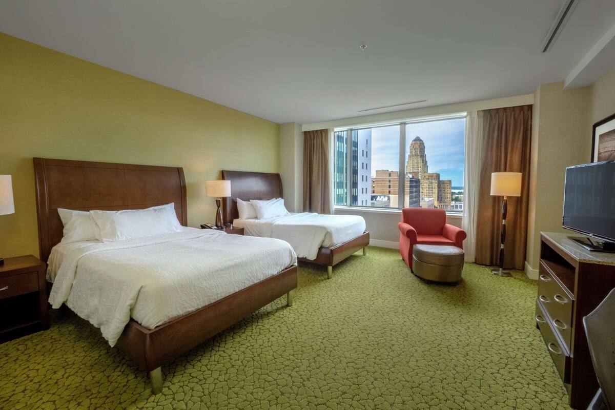 Une chambre d’hôtel avec deux lits, une télévision, une chaise et une vue sur le paysage urbain.