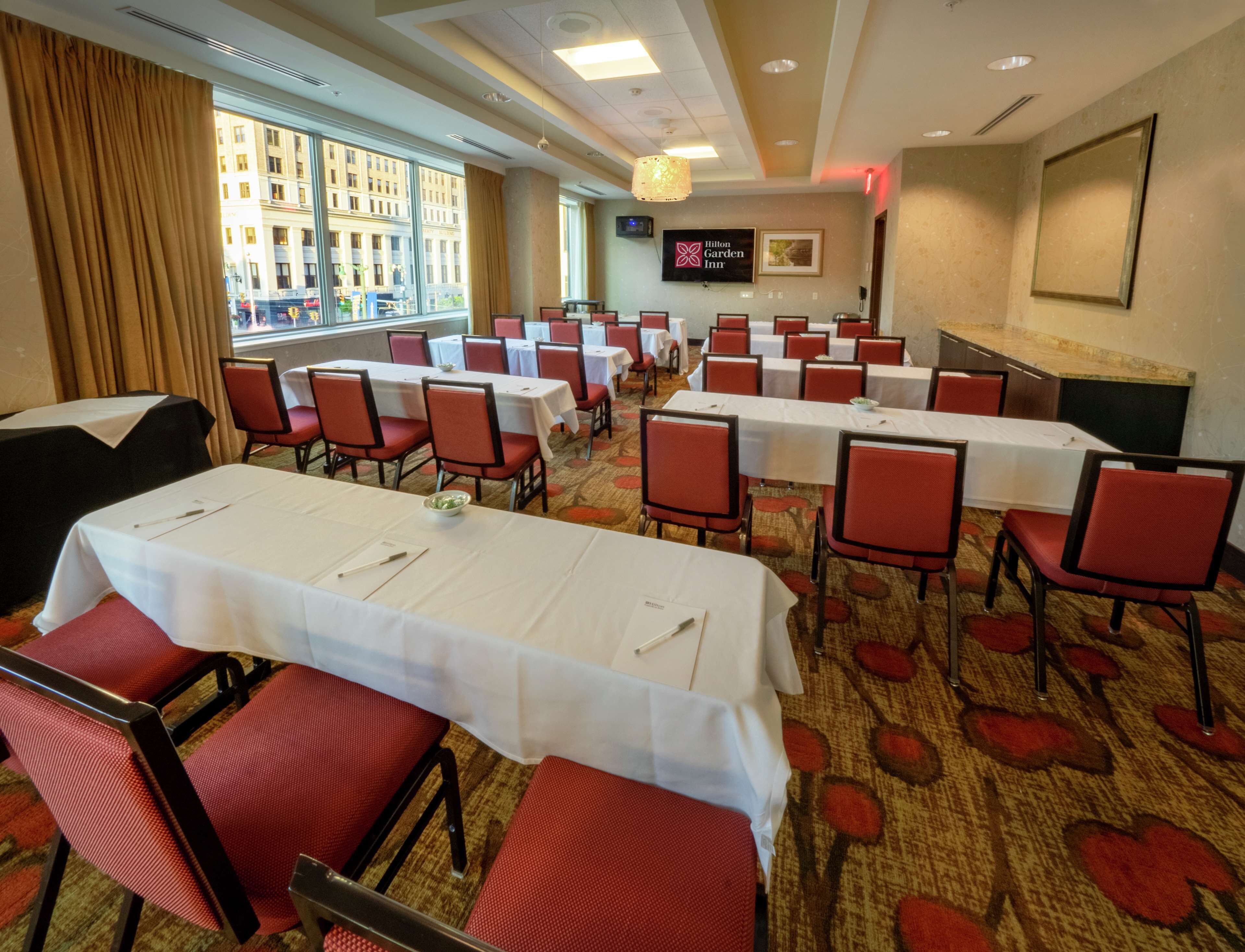 A hotel conference room with tables and chairs, a television, and a view of the city through large windows.