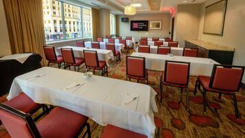 A hotel conference room with tables and chairs, a television, and a view of the city through large windows.