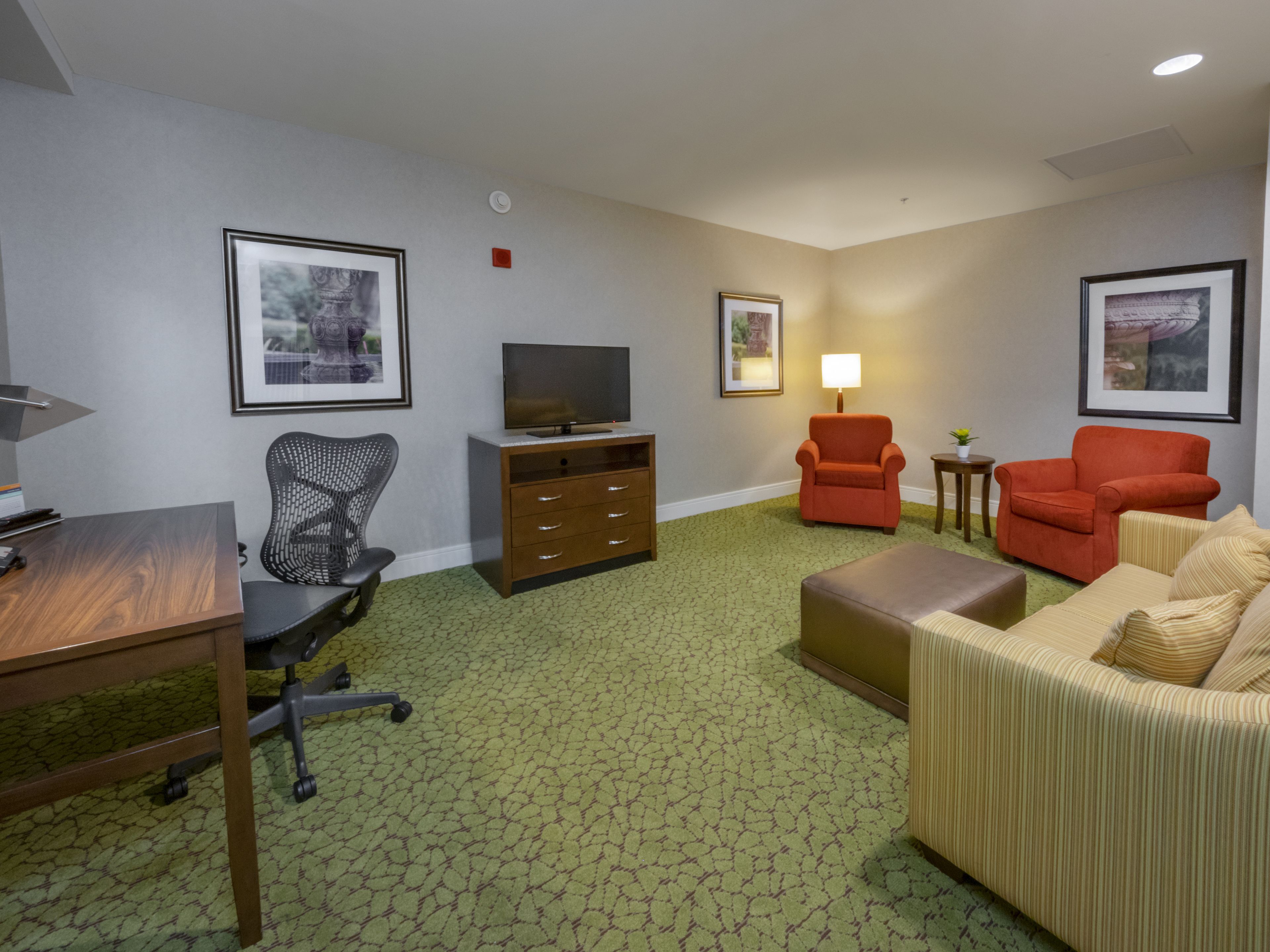 A hotel room with a wooden desk, a black office chair, a brown sofa, a red armchair, a television on a wooden cabinet, and two framed pictures on the walls.