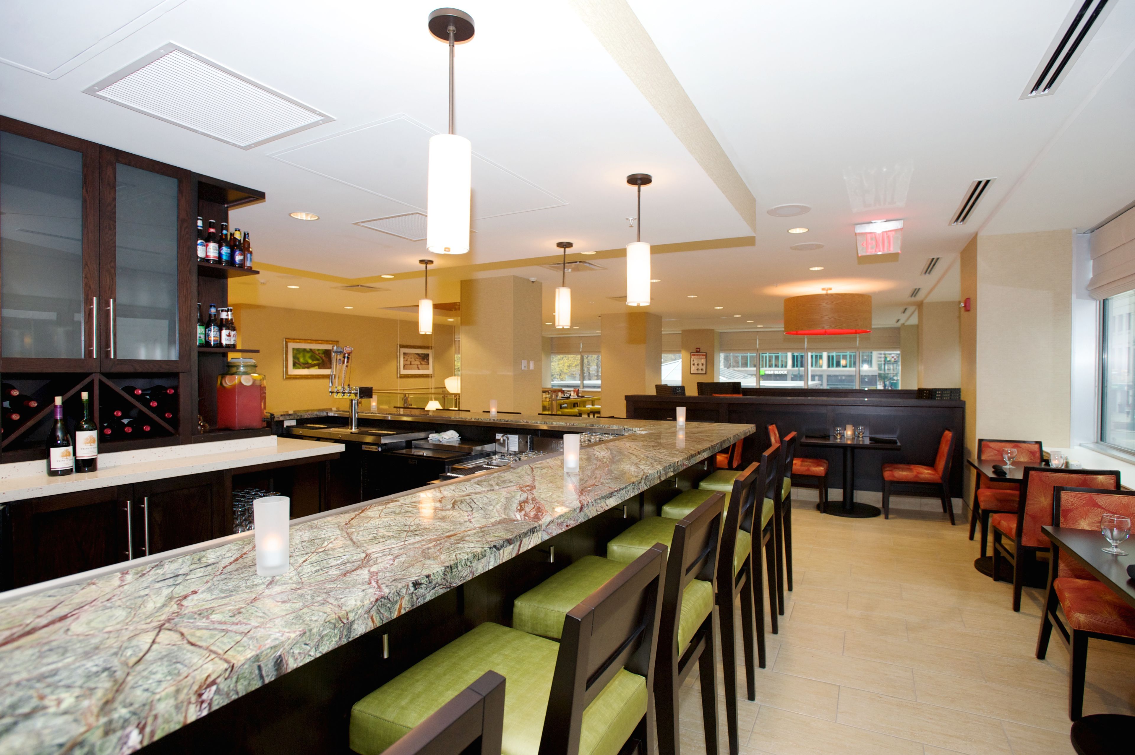 A restaurant with a marble bar, green cushioned bar stools, and a seating area with wooden tables and chairs.
