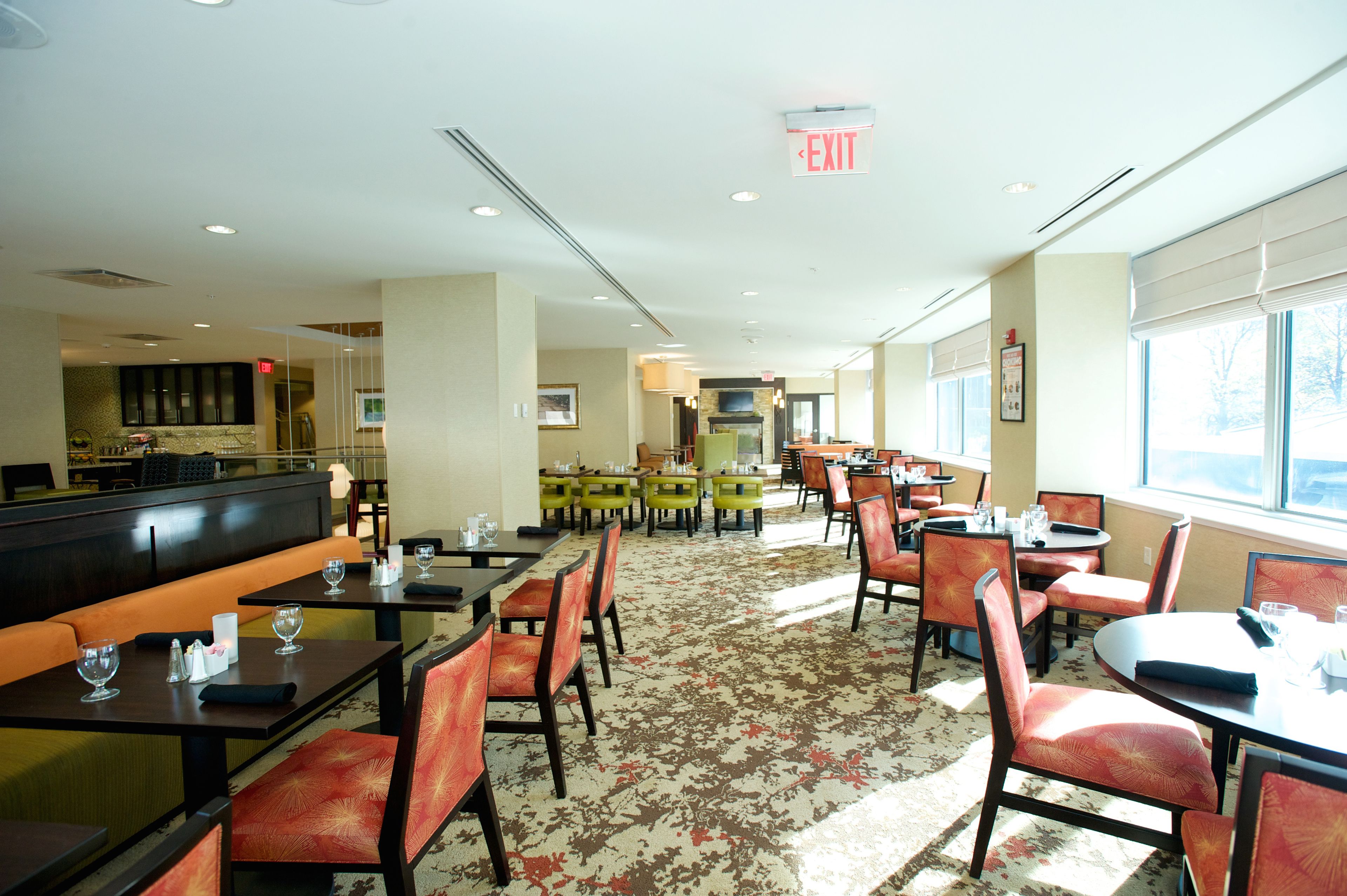 A hotel dining area with multiple tables and chairs, a kitchen in the background, and an exit sign on the ceiling.