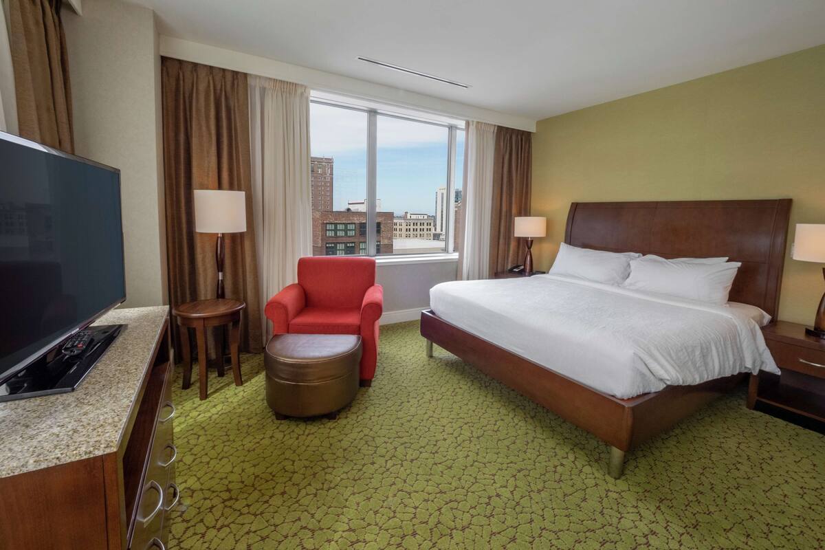 Une chambre d’hôtel avec un grand lit, une télévision, un fauteuil rouge, un petit pouf et une vue sur la ville par la fenêtre.