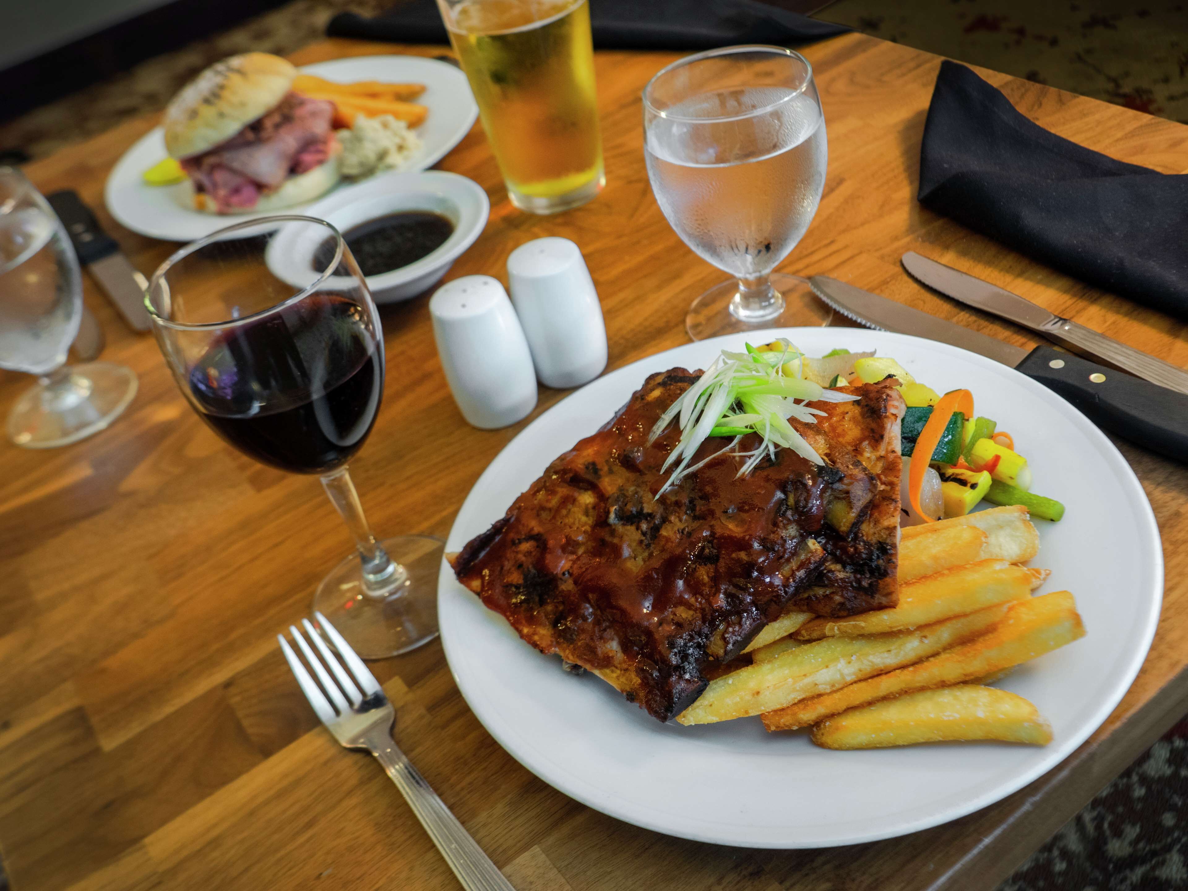 A dining table with a plate of ribs, fries, and vegetables, a glass of red wine, and a beer glass.