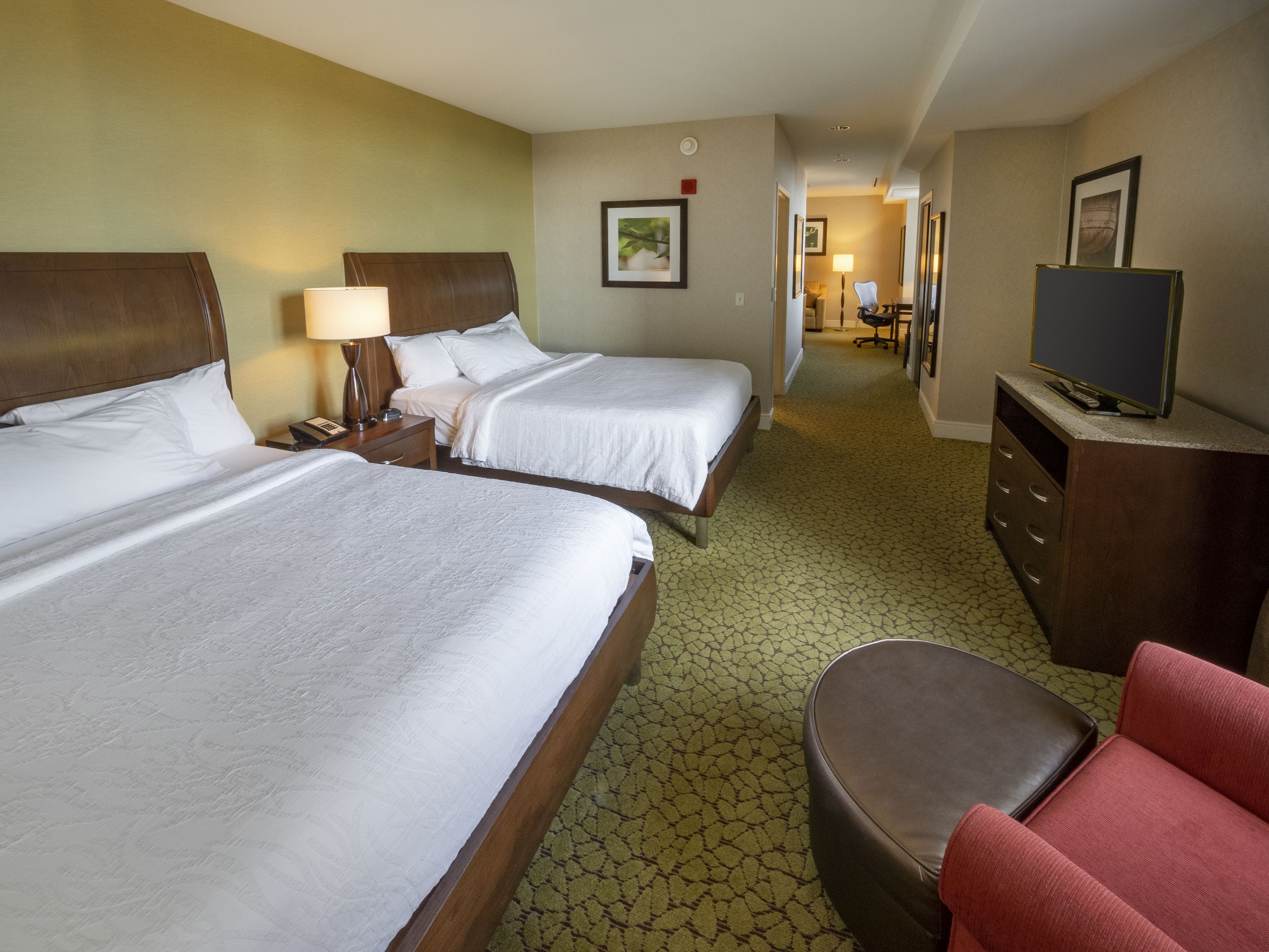 A hotel room with two beds, a desk, a chair, a TV, and a hallway leading to another room.