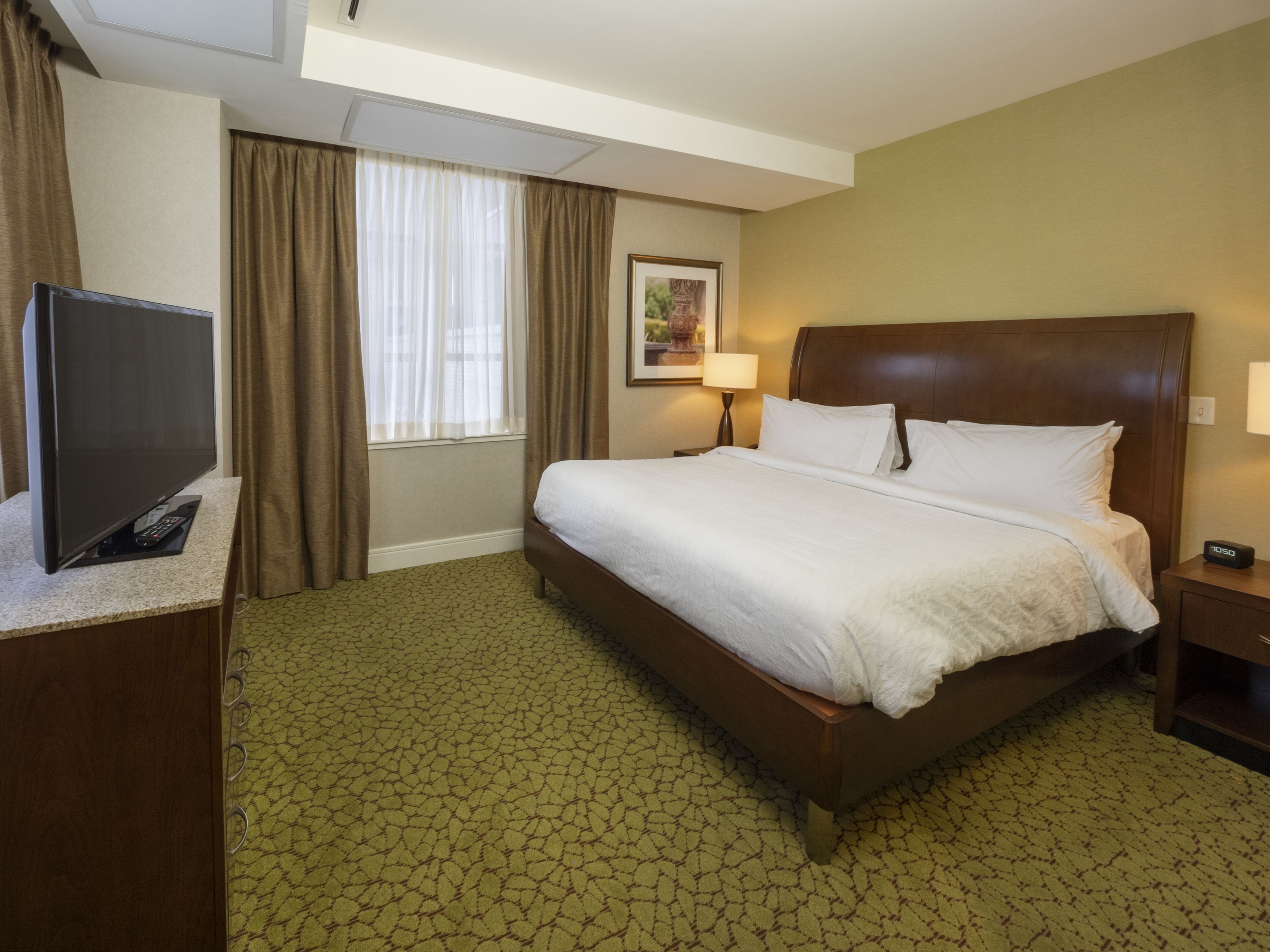Ein Hotelzimmer mit einem großen Bett, einem Fernseher auf einem Tisch, einem Schreibtisch und einem Fenster mit Vorhängen.