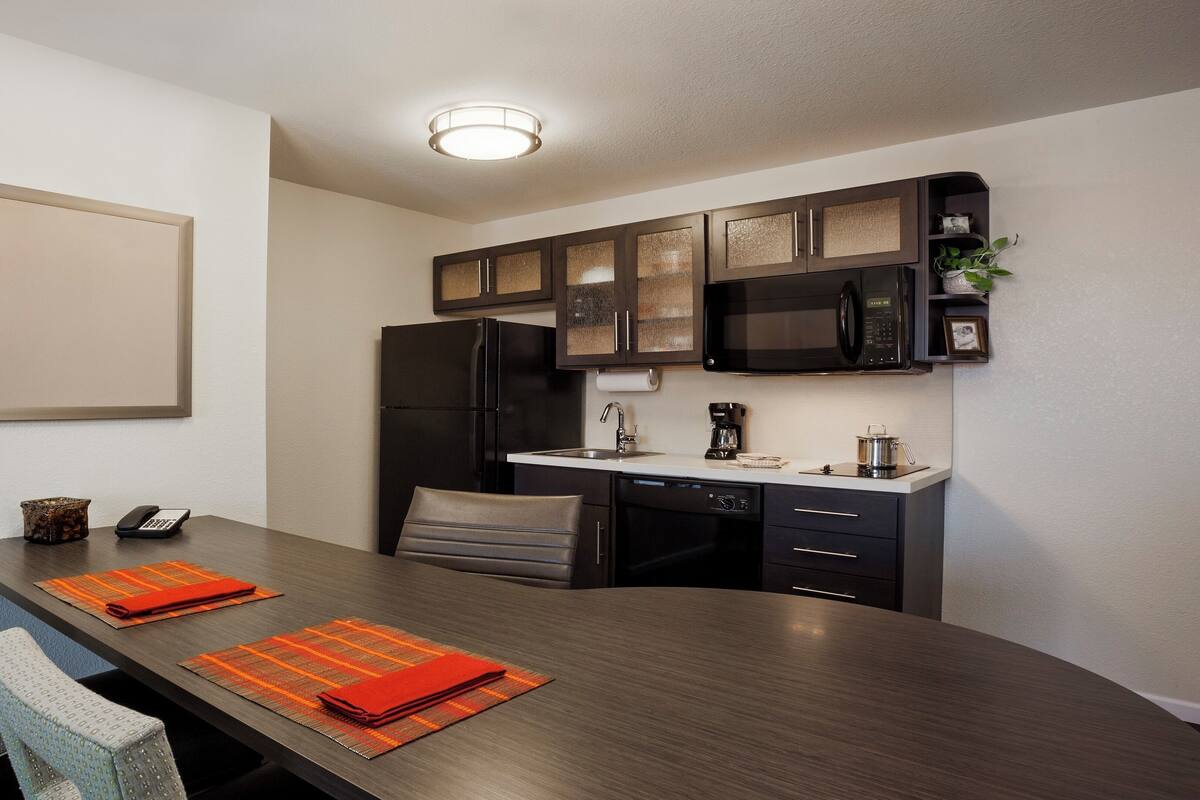 Una cocina moderna con armarios oscuros, un refrigerador negro, un microondas y una cafetera. Hay una mesa de comedor con dos sillas y manteles individuales naranjas.