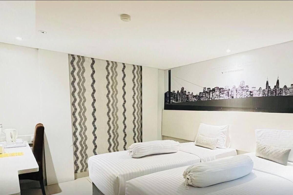 A hotel room with two twin beds, a cityscape wall art, a patterned curtain, and a desk with a chair.