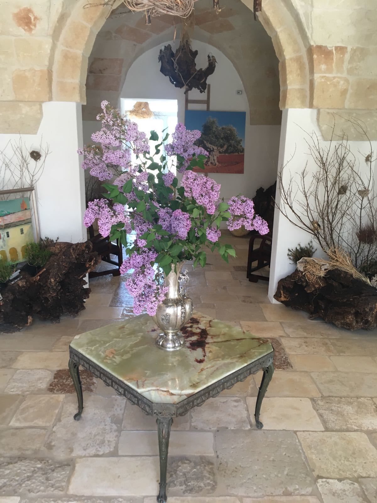 Une table en marbre surmontée d’un vase en argent contenant des fleurs violettes, le tout situé dans un couloir à arcades.