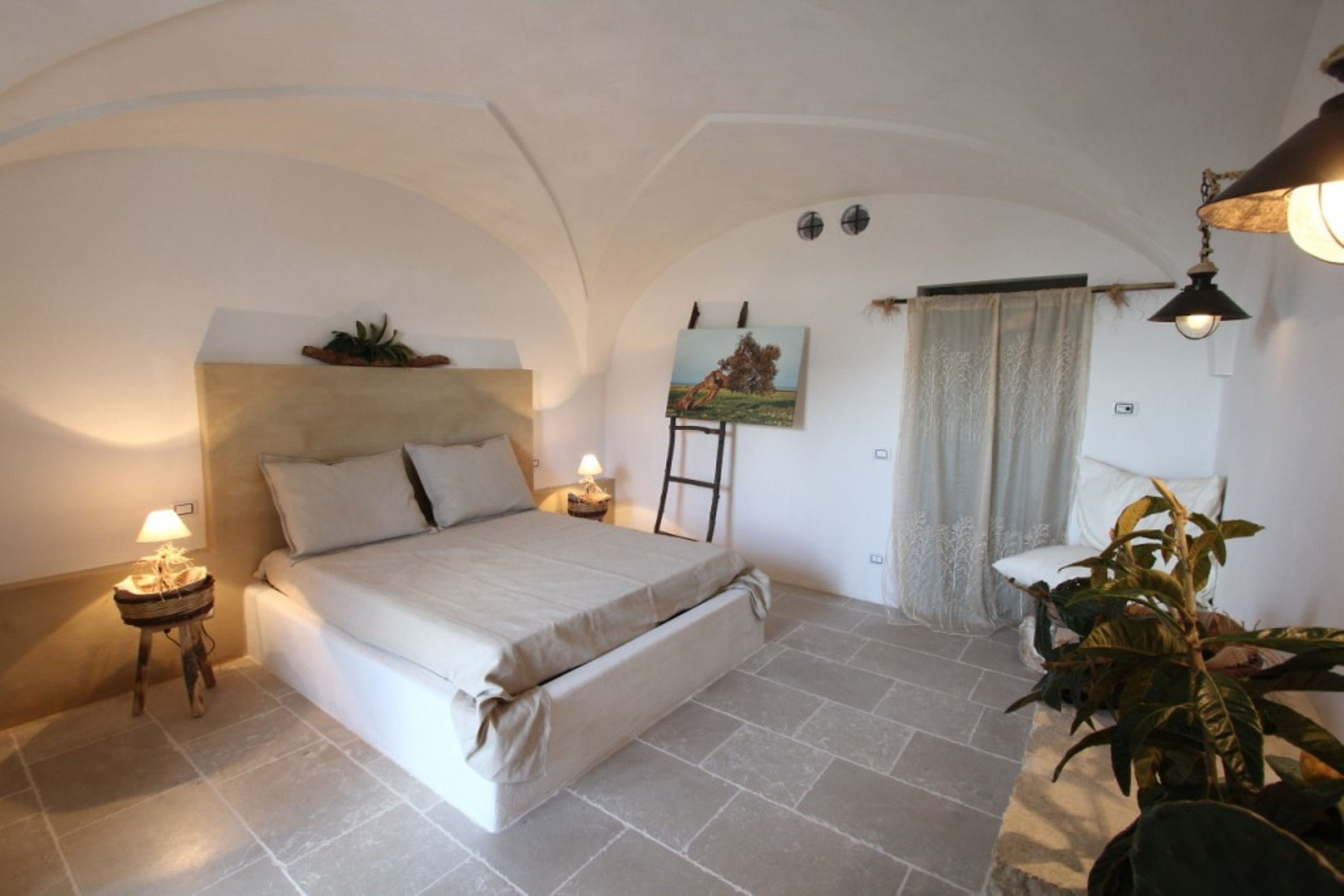 A bedroom with a bed, bedside table, lamp, painting, and curtains.