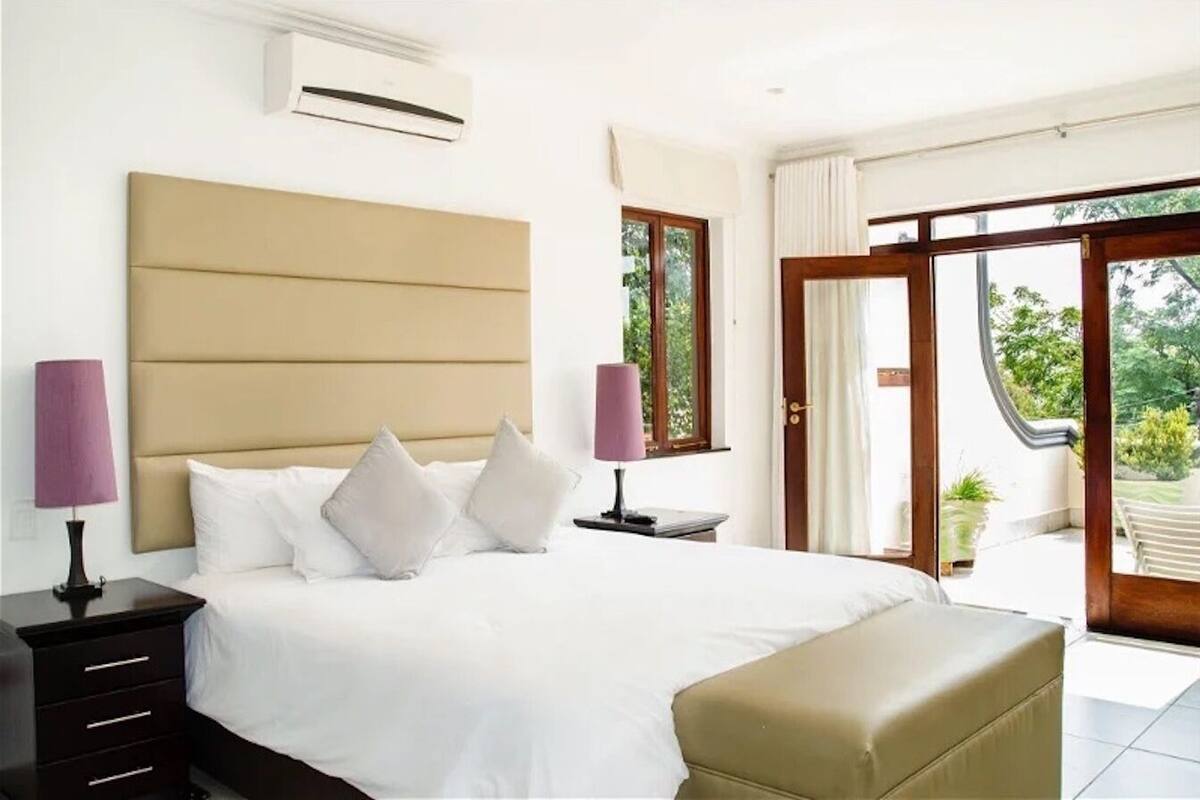 Quarto com cama grande, mesas de cabeceira, um banco e vista para o exterior através de uma porta de correr de vidro.