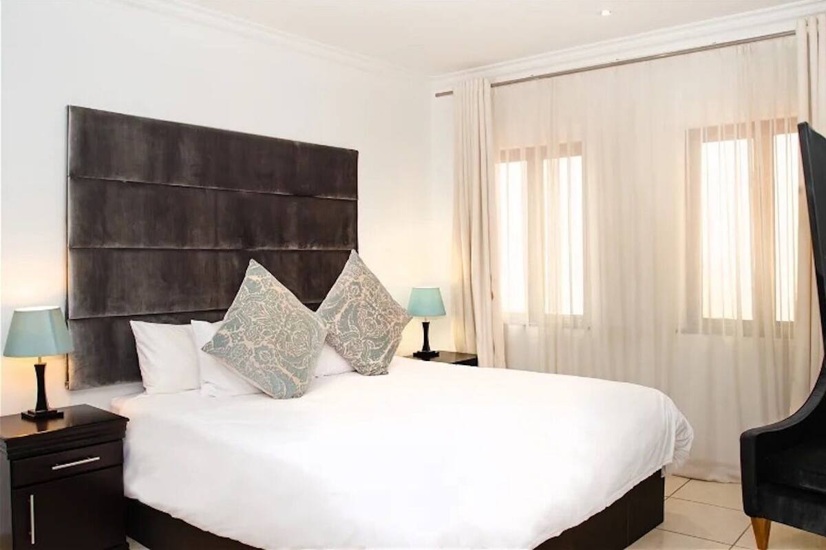 Quarto de hotel com uma cama grande, duas mesas de cabeceira com luminárias, um painel de cabeceira escuro e uma grande janela com cortinas.