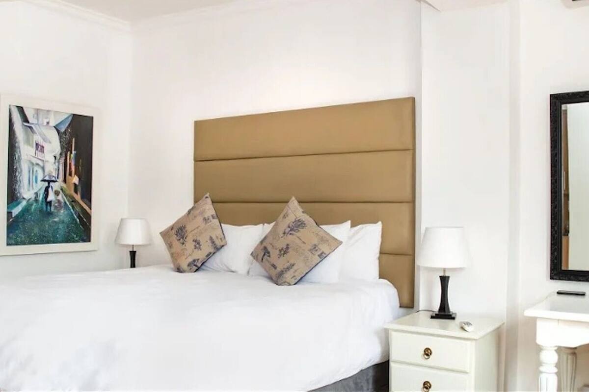 Quarto de hotel com uma cama, duas mesas de cabeceira, um criado-mudo, um espelho e um quadro na parede.
