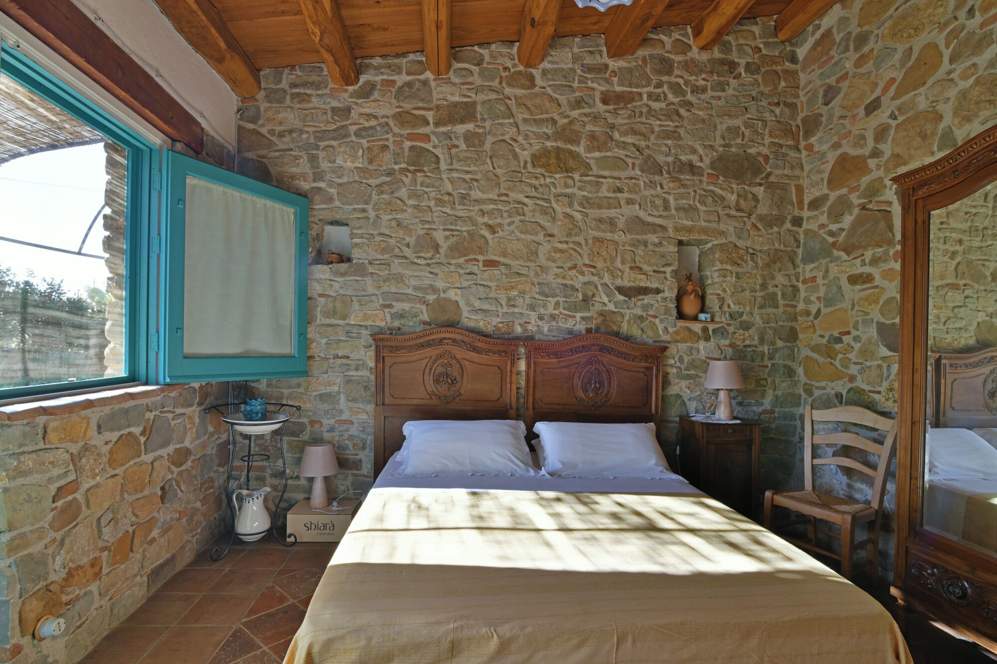 A bedroom with a stone wall, a wooden ceiling, a bed with white linens, a bedside table with a lamp, and a window with a blue frame.