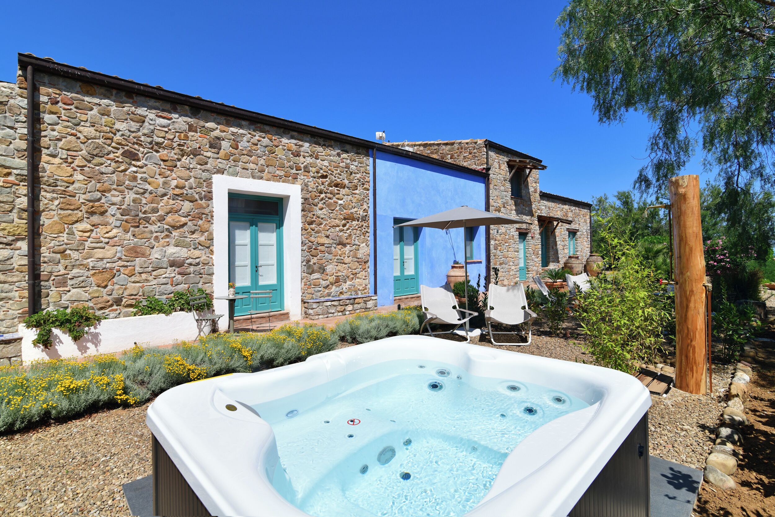A stone building with a hot tub in the foreground, outdoor seating, and a clear blue sky.