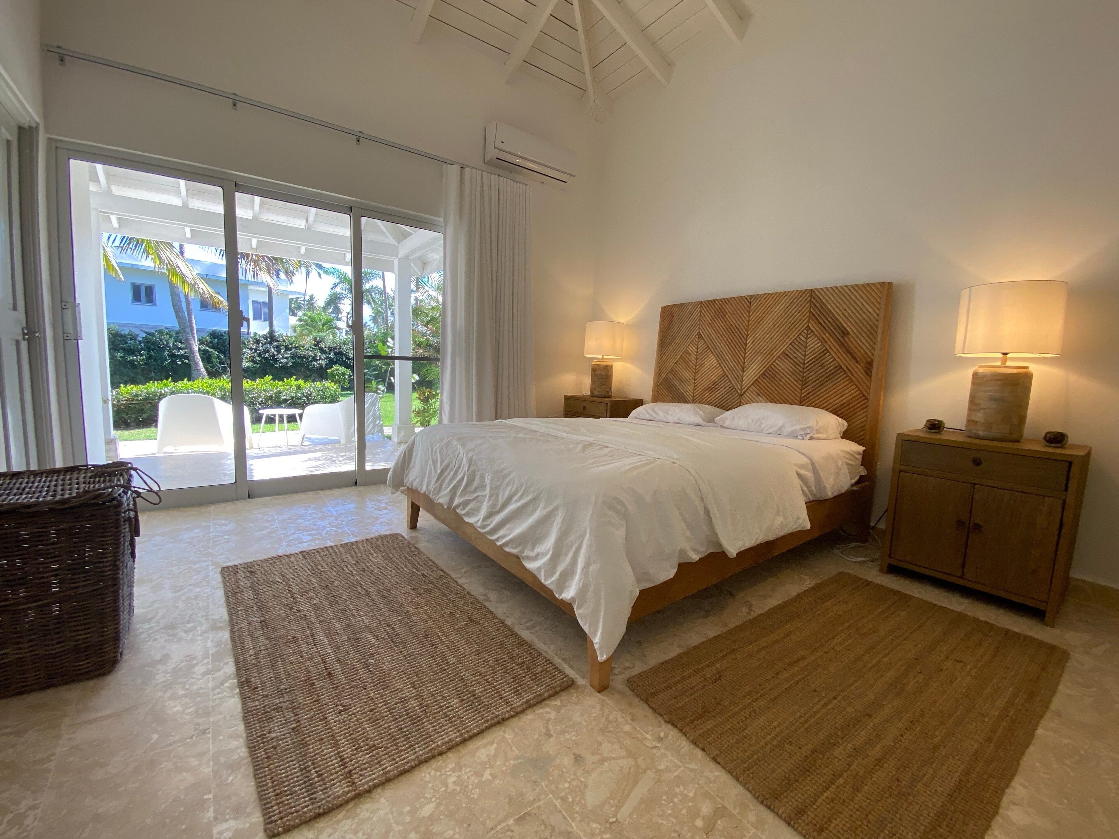 A bedroom with a large bed, two bedside lamps, a wooden nightstand, a wicker chair, and a view of a garden through a sliding glass door.