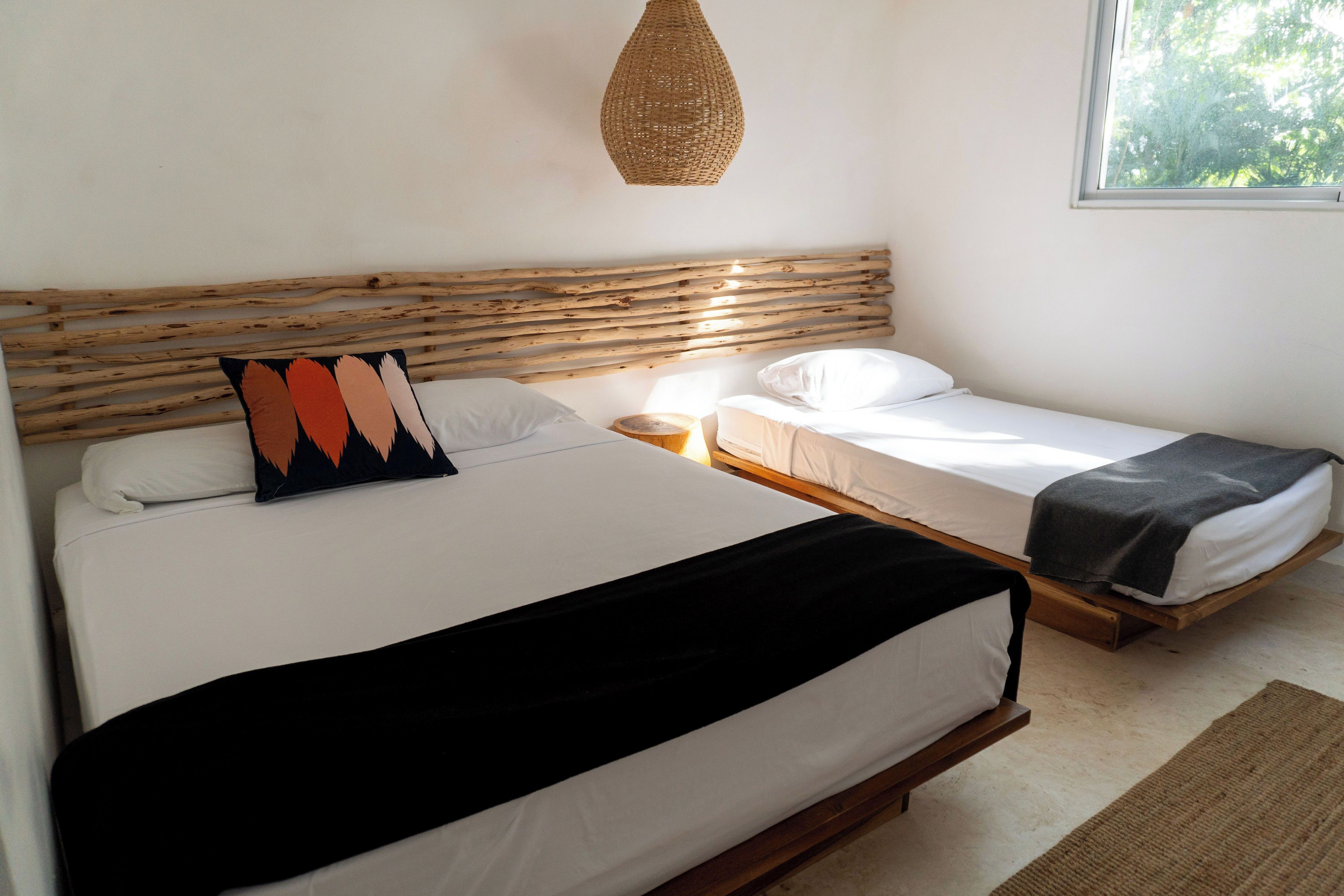 A bedroom with two beds, a woven light fixture, and a window with a view of greenery.
