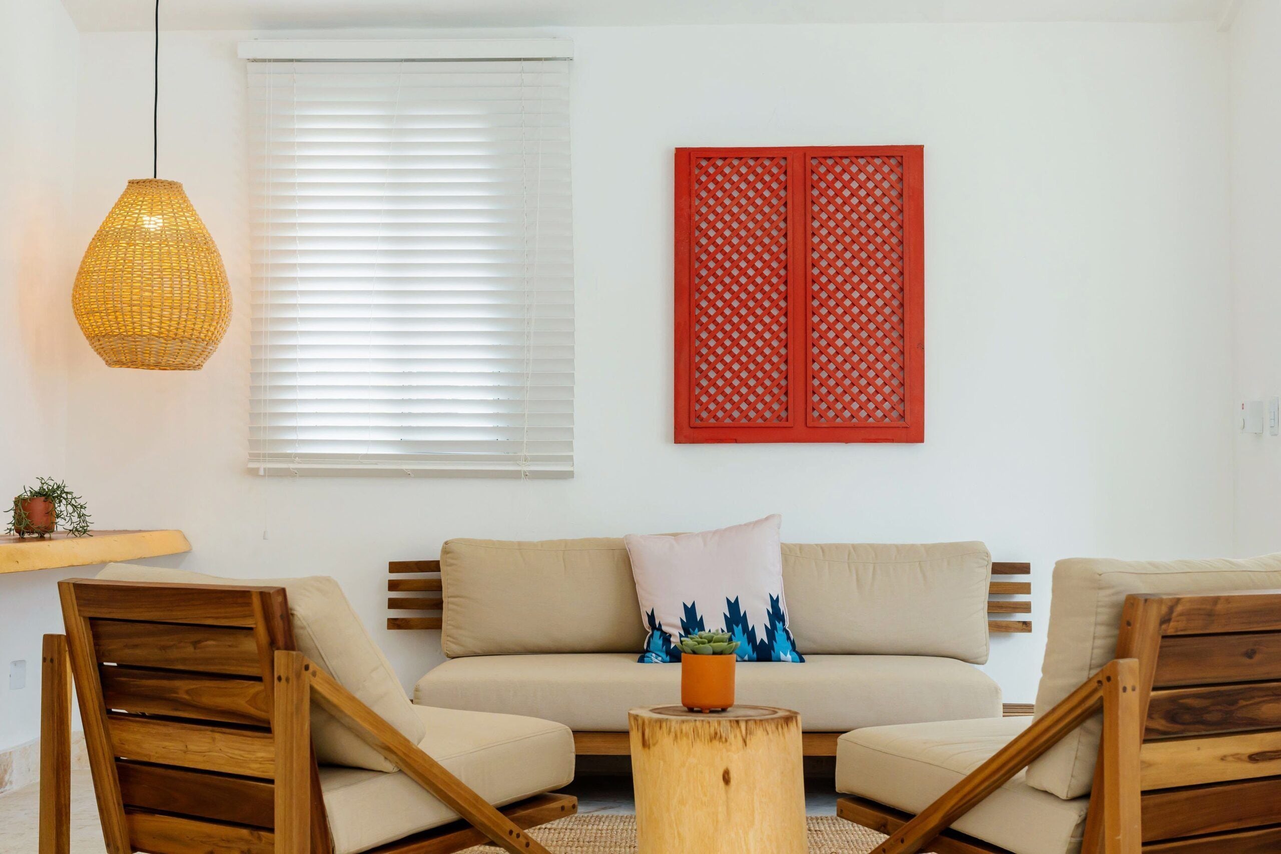 A living room with a sofa, two wooden chairs, a woven pendant light, a red decorative panel, and a small plant on a wooden stool.