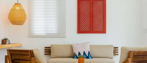 A living room with a sofa, two wooden chairs, a woven pendant light, a red decorative panel, and a small plant on a wooden stool.