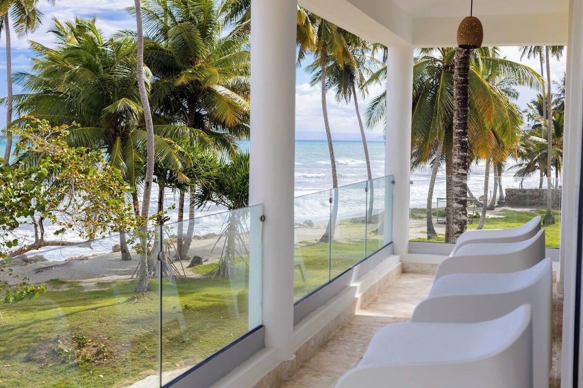 A balcony with a glass railing overlooking a beach with palm trees and the ocean.