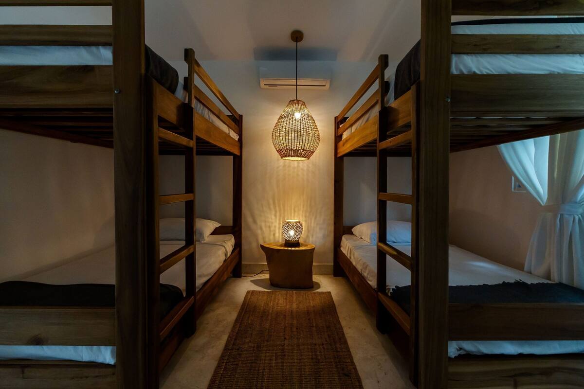 A room with bunk beds, a central hanging light fixture, and a small side table.