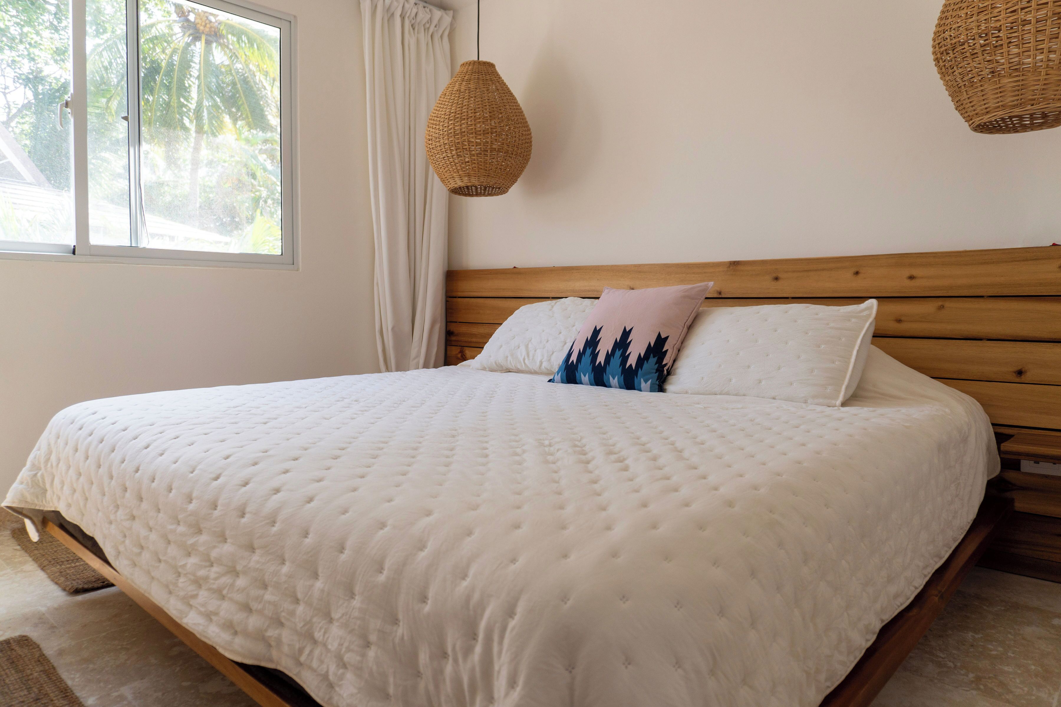Un lit bien fait, recouvert d’une couette blanche, avec une tête de lit en bois et une fenêtre agrémentée de rideaux.