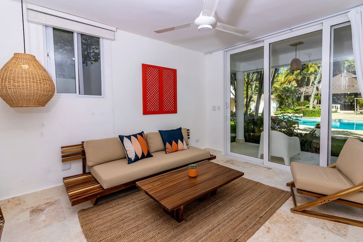 A modern living room with a sofa, a coffee table, a chair, a large window with a view of a pool, and a ceiling fan.