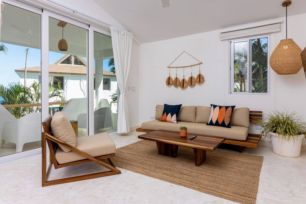A modern living room with a sofa, a wooden coffee table, and a chair. There is a large window with a view of palm trees and a building outside.