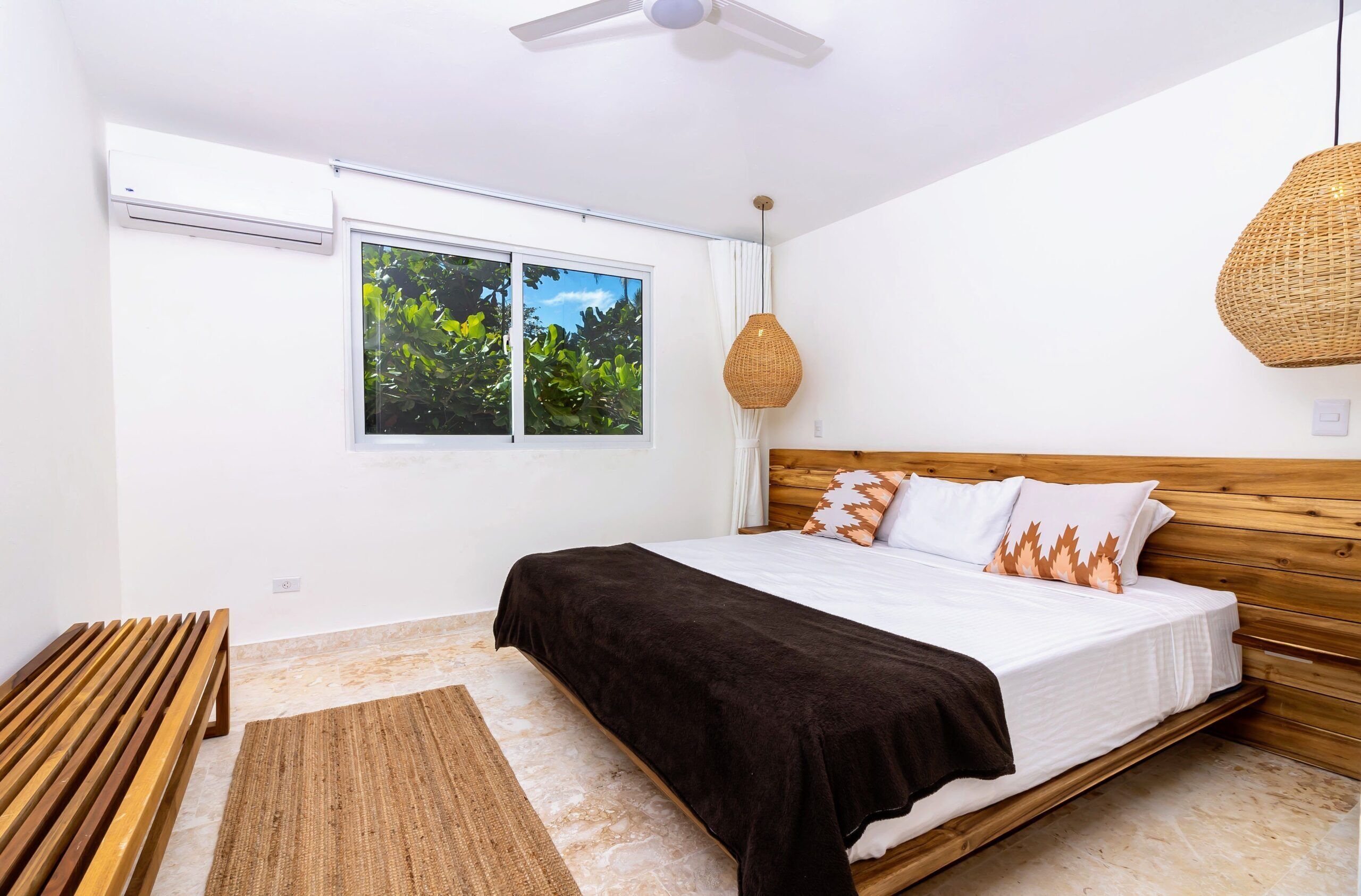 A bedroom with a bed, a wooden bench, a woven light fixture, and a window with a view of greenery.