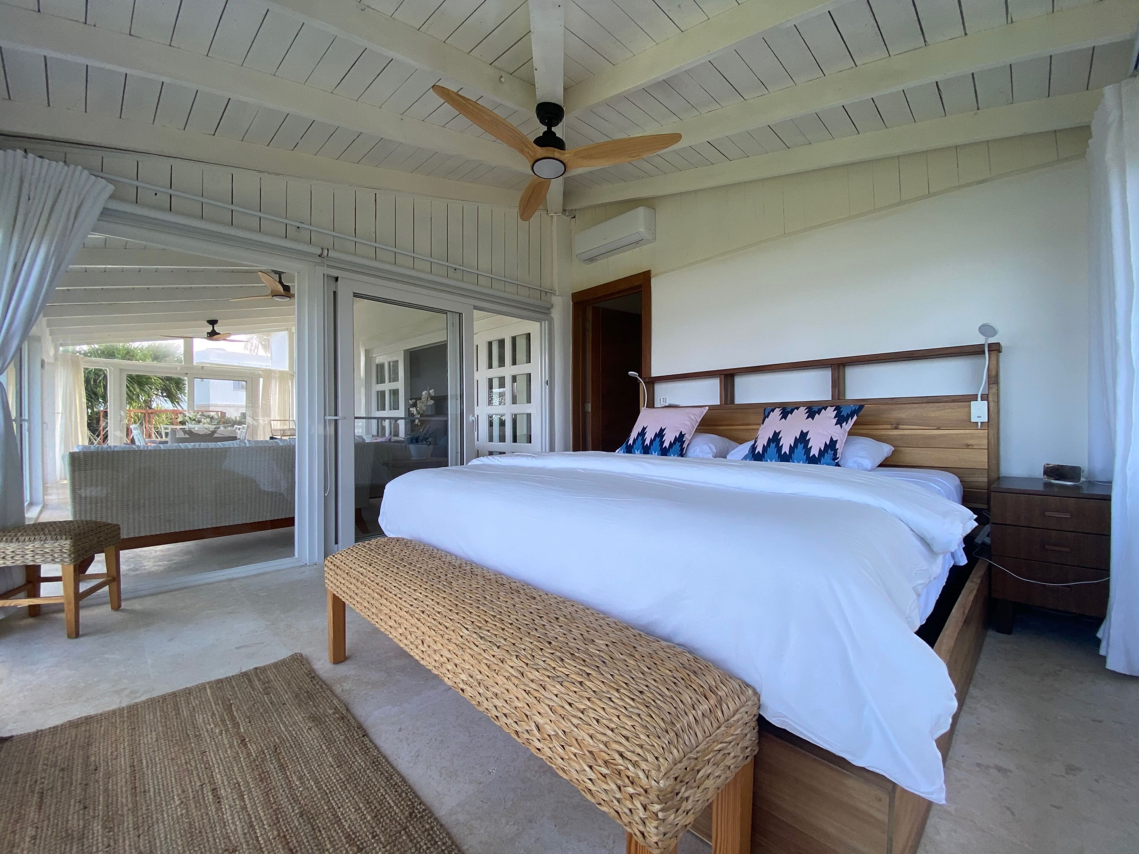 A bedroom with a large bed, a wooden headboard, a bedside table, a bench, a ceiling fan, and a sliding glass door leading to an outdoor area.