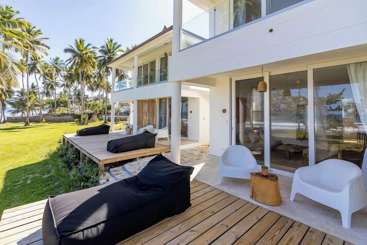 A modern two-story house with a deck, outdoor seating, and a view of palm trees.