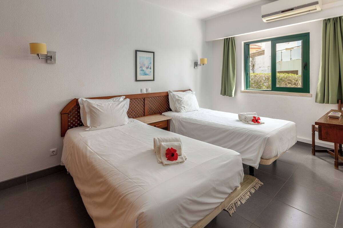 Habitación de hotel con dos camas, una mesita, una mesita de noche, un cuadro enmarcado en la pared y una ventana con cortinas.