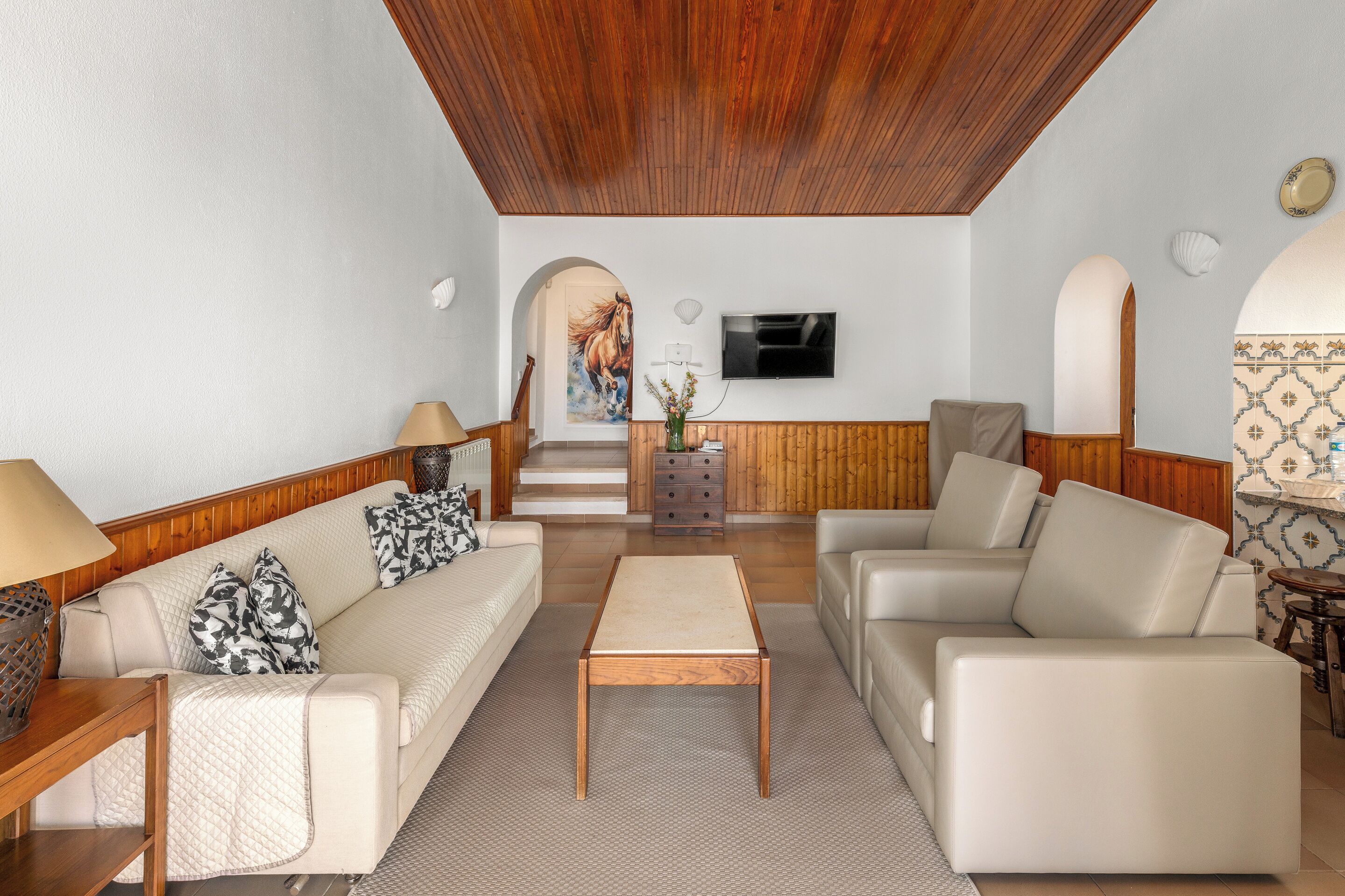 A living room with a wooden ceiling, a TV mounted on the wall, a sofa, a coffee table, and a patterned rug.