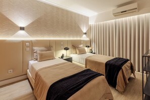 A hotel room with two beds, a large window with blinds, an air conditioning unit, and a bedside table with a lamp.