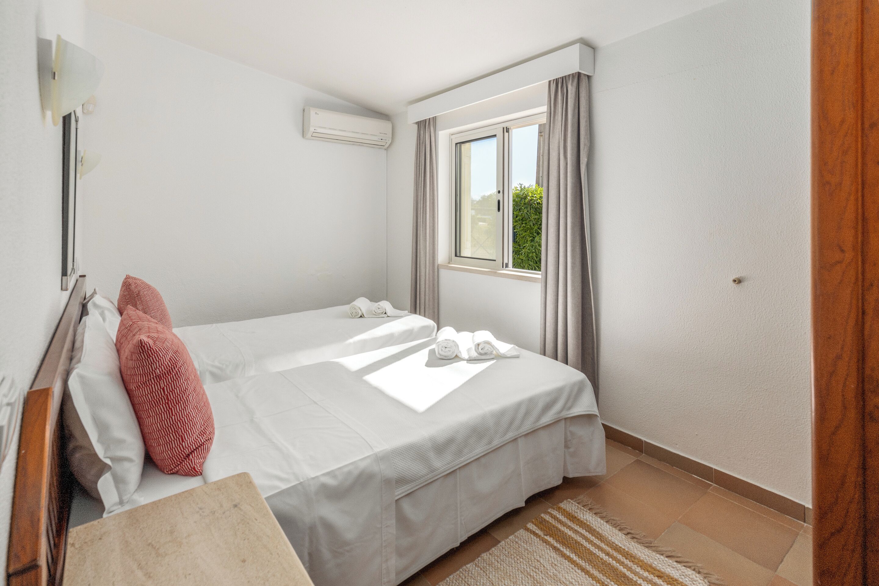 A hotel room with two beds, a window with curtains, an air conditioning unit, and a wooden door.